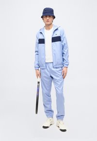 Νεαρός άνδρας με ανοιχτό μπλε φόρμα, ναυτικό καπέλο τύπου κάδο και λευκά αθλητικά παπούτσια που κρατάει ρακέτα τένις, στέκεται μπροστά σε ένα απλό φωτεινό φόντο.