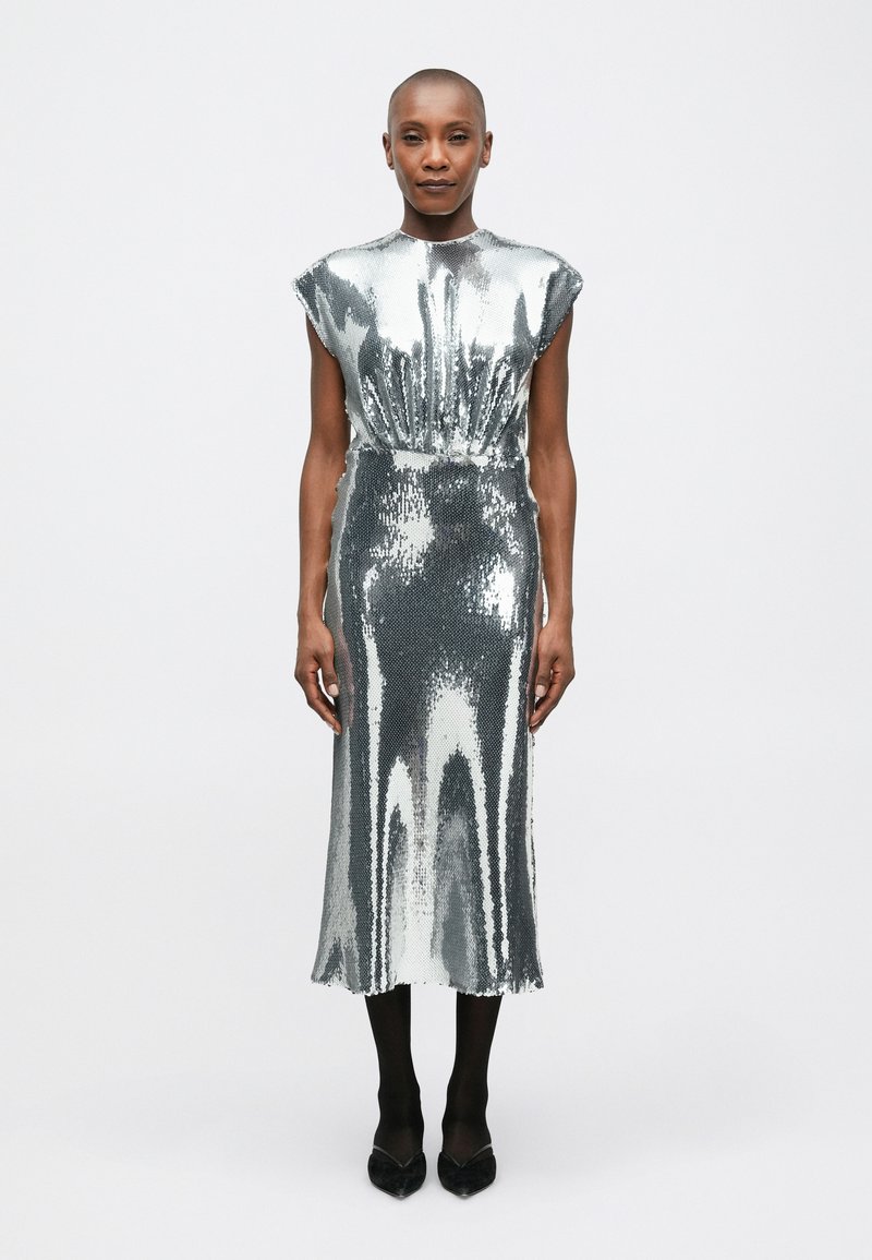 Cilvēks stāv taisni, ģērbies bez piedurknēm sudraba spigulīša kleitā, melnās zeķbiksēs un melnās norādītās kurpēs uz vienkārša fona.