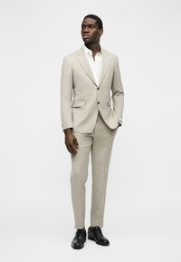 Άνδρας όρθιος, ντυμένος με μπεζ κοστούμι, λευκό πουκάμισο και μαύρα επίσημα παπούτσια, με τα χέρια χαλαρά, κοιτάζοντας ελαφρώς προς το πλάι.