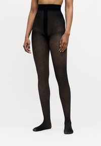 Person wearing sheer black tights with small polka dots, standing barefoot against a plain white background.