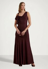 Vestido bordô de cetim, de um ombro, apresentando um corpete drapeado e um detalhe floral. Modelo segura uma clutch a condizer, combinada com sapatos de salto aberto.