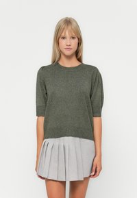 Pull en maille vert olive avec des manches courtes bouffantes, associé à une jupe plissée gris clair. Texture douce, design décontracté et coupe relax.