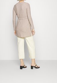 Beige, gerippter Langcardigan mit Bindegürtel, kombiniert mit cremefarbenen, weiten Hosen und schwarzen Blockabsatzsandalen. Seitenschlitze am Cardigan.
