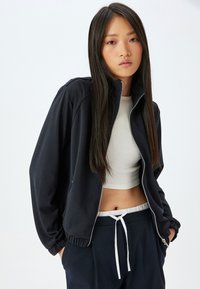 Svart zip-up jacka med elastiska ärmslut, kombinerad med en ljusbeige kort topp. Texturerad tyg i avslappnad passform, bärs med mörka byxor.
