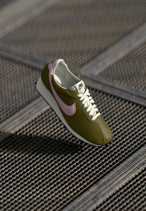 Nike tenisky, olivově zelená látka s ribbed texturou, růžové akcenty, bílé šňůrky a bílá podrážka, které ukazují retro sportovní design.