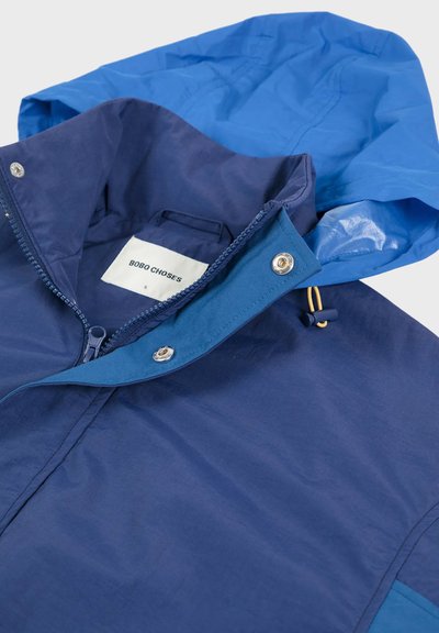 Navy blue rain jacket with a blue hood, featuring a front zipper, snap buttons, and a label inside. Smooth texture and lightweight material.