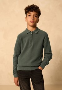 Zelený pletený svetr na zip s límcem, žebrovanou texturou a dlouhými rukávy. Nošený s tmavými džíny; ležérní design zaměřený na jednoduchost.