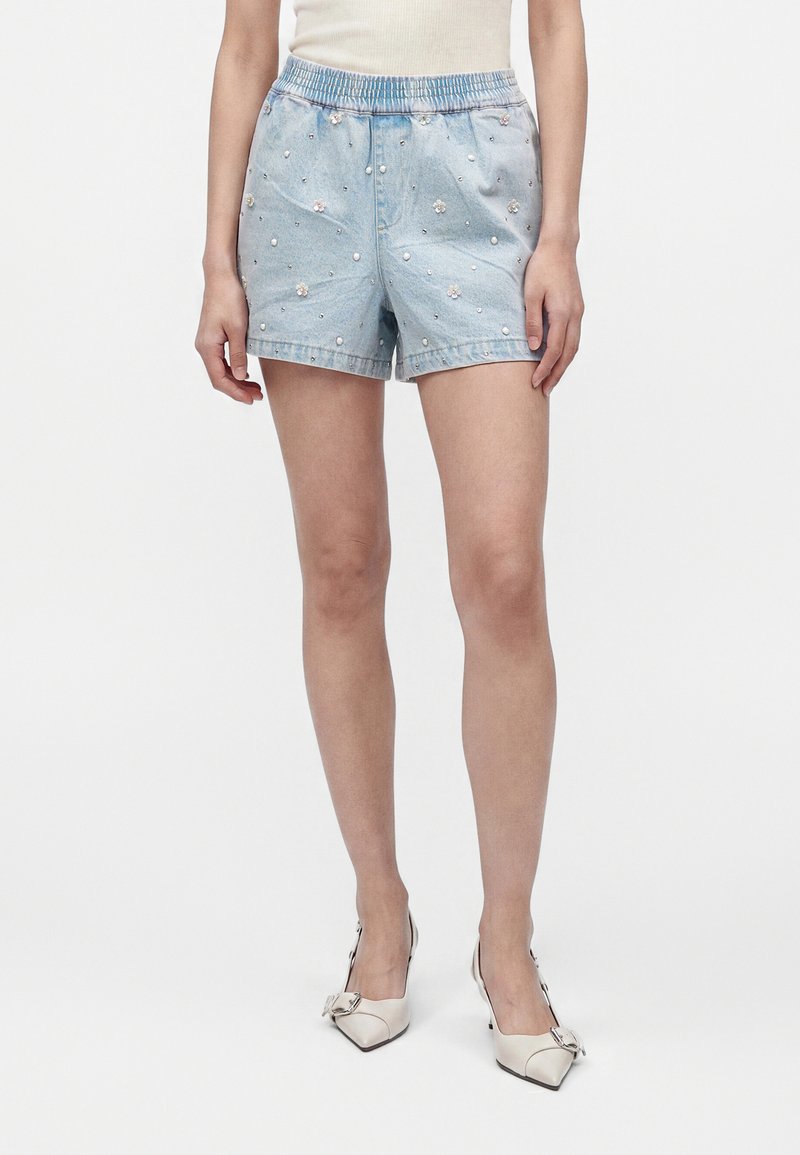 Femme portant un short en jean bleu clair orné de perles et de fleurs, associé à des chaussures crème à talons bas avec boucles.