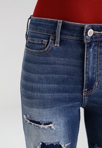 Tmavě modré džíny z denimu se podlepenými detaily, s jemným gradientem, roztrhanými koleny, designem s pěti kapsami a viditelnými švy.