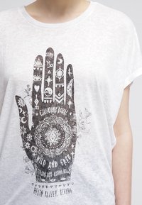 Camiseta blanca con un gran gráfico negro de una mano que presenta varios símbolos, patrones y texto en un estilo desgastado. Corte corto y holgado.