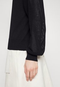 Černý pletený svetr s texturovanými rukávy a žebrovanými manžetami, v kombinaci se světle zbarvenou plisovanou sukní. Tkanina vypadá měkce a útulně.