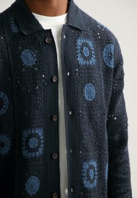 Cardigan tricotat de culoare albastru marin cu modele florale albastre intricate. Are un design cu guler, închidere cu nasturi și material texturat.