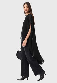 Haut noir ample en tissu transparent avec des fentes latérales, associé à un jean noir taille haute et des escarpins noirs pointus.