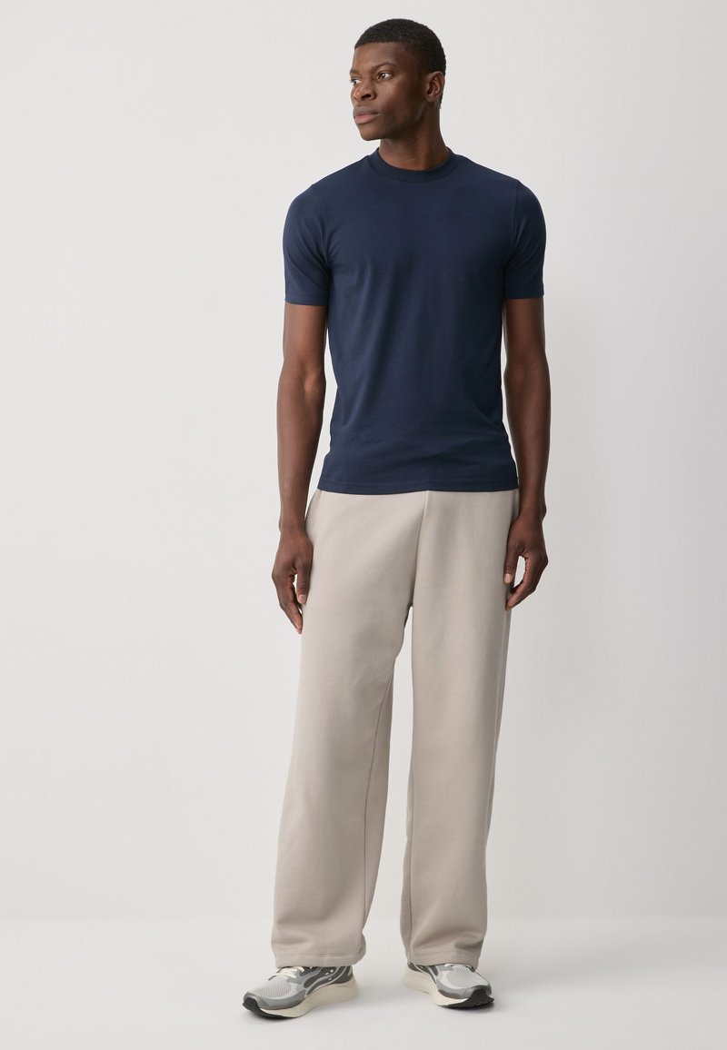 Homme debout regardant vers la gauche, portant un t-shirt bleu marine à manches courtes, un pantalon beige à jambes larges et des chaussures de sport grises sur un fond uni.