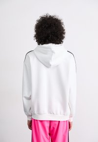 Sweatshirt branco com capuz feito de um tecido macio, apresentando riscas pretas nos ombros, punhos e cós canelados, usado com calças de treino cor-de-rosa.