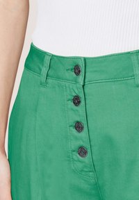 Grüne Jeans mit hohem Bund, fünf schwarzen Metallknöpfen und sichtbaren Nähdetails. Die Textur ist glatt mit einem leichten Glanz.