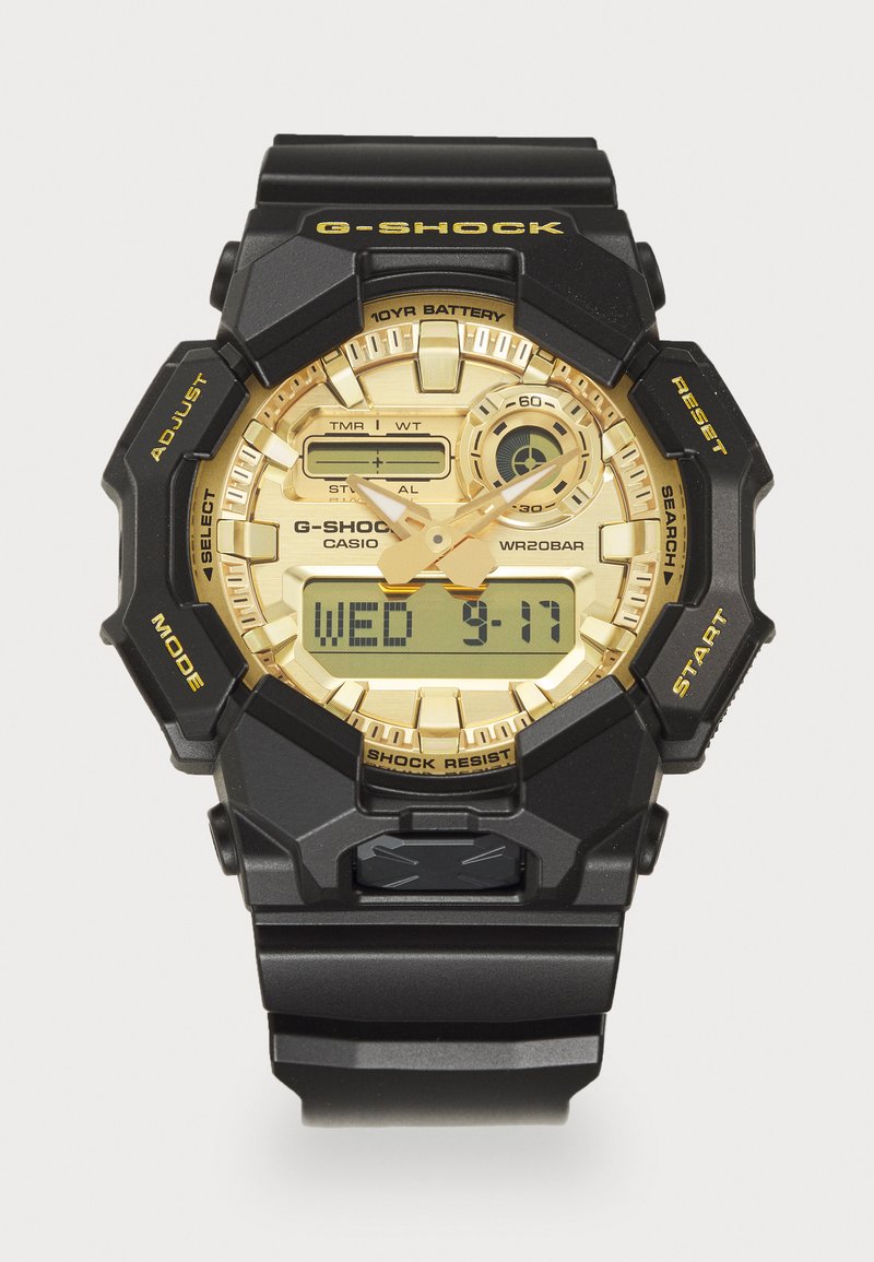 Hodinky G-SHOCK s čiernym remienkom z živice a zlatým ciferníkom. Obsahujú digitálny displej, chronograf a viacero tlačidiel na funkcie.