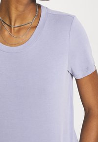 Ljuslila t-shirt med rund halsringning, korta ärmar, slät bomullstextur och subtila sömnadsdetaljer. Silvrig lagerhalsband synligt.