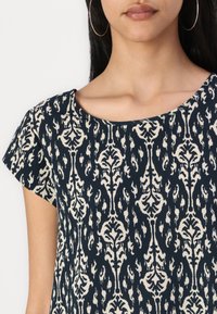 Marineblå topp med kremfarget blomsterdesign, rund hals, og korte cap sleeves. Stoffet ser mykt ut med en lett tekstur.