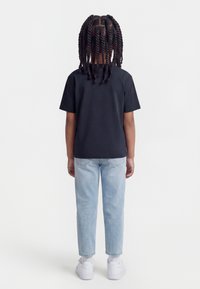 Παιδί με μακριά στριφτά μαλλιά στέκεται με την πλάτη γυρισμένη, φορώντας μαύρο t-shirt, ανοιχτό μπλε τζιν, λευκές κάλτσες και λευκά αθλητικά παπούτσια, μπροστά σε λευκό φόντο.