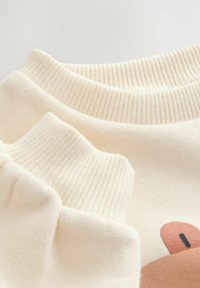 Pull couleur crème avec poignets et col côtelés, texture douce, et un imprimé graphique d'un personnage de dessin animé sur le devant.