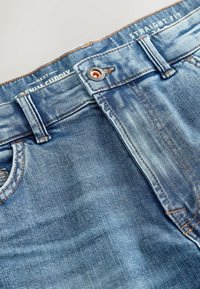 Jasnoniebieskie dżinsy z denimu o prostym kroju, wyposażone w zapięcie na guzik, miedziane okucia oraz widoczne przeszycia wzdłuż talii.