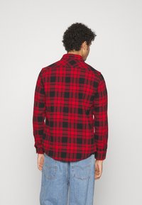 Camisa de franela a cuadros en rojo y negro con mangas largas y puños de botón. Corte entallado, con cuello puntiagudo y dobladillo recto.