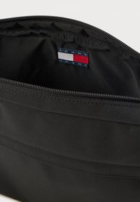 Indvendig i en sort nylon tote taske med lynlås. Har et "TOMMY JEANS" mærke med blå, rød og hvid farveakcent. Glat tekstur.