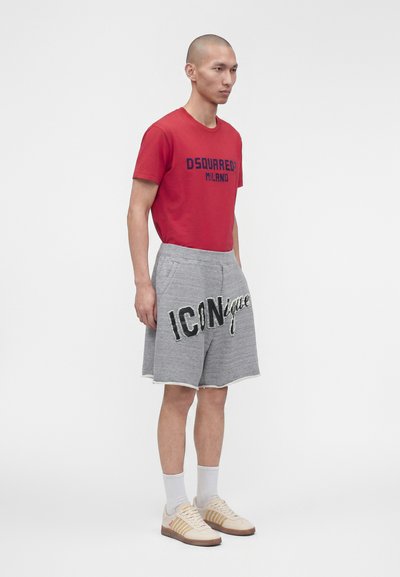 Άνδρας μοντέλο που φοράει κόκκινο t-shirt Dsquared2 Milano, γκρι σορτς με το κείμενο "ICONique", λευκές κάλτσες και μπεζ αθλητικά παπούτσια, στέκεται μπροστά σε λευκό φόντο.
