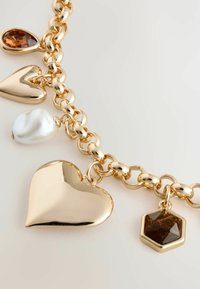 Guldkædearmbånd med hjerte, tåreformede og hexagonale charms, en med brun sten og en hvid perle som accent. Glat, skinnende finish.