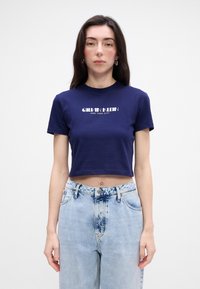 Jūrinės spalvos trumpa marškinė su baltais užrašais "Calvin Klein New York City", apvaliu kaklu, trumpomis rankovėmis, derinami su šviesiai mėlynomis džinsinėmis kelnėmis.