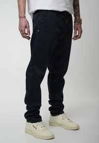 Pantaloni tapered blu navy con vita regolabile, tasche laterali e posteriori, abbinati a sneakers crema. Il tessuto ha una texture liscia.