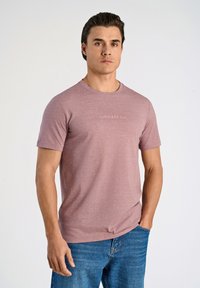 Ljusrosa kortärmad t-shirt med rund halsringning och subtil logotyp i mitten, i kombination med blå denimjeans. Enkel, avslappnad design.