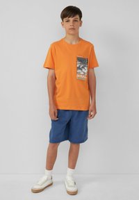 Oranje katoenen t-shirt met een zwart-wit grafisch ontwerp op de zak, gecombineerd met blauwe shorts en witte schoenen met beige zolen.