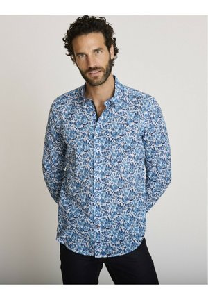 Homme aux cheveux bouclés portant une chemise bleue à manches longues à motifs floraux, debout devant un fond clair uni.