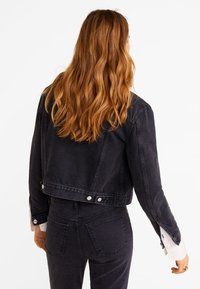 Mujer con cabello rojo largo y ondulado, vistiendo una chaqueta de mezclilla negra y jeans negros, vista desde atrás contra un fondo blanco.