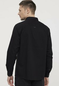 Zwarte lange mouwen shirt met een kraag met knopen, zichtbare naaddetails, een lus aan de achterkant en met knopen dichtgeknoopte manchetten.