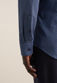 Camicia navy con un motivo geometrico blu, dotata di polsino con bottoni e texture liscia, mostrata sul braccio di una persona contro uno sfondo neutro.