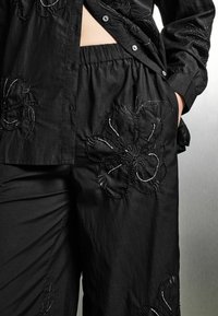 Schwarzes Baumwollhemd mit Knopfverschluss, verziert mit strukturierten floralen Stickereien in weißen Fäden. Passende schwarze Hose mit ähnlichen floralen Akzenten.