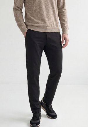 Mann, der schwarze Hose, schwarze Turnschuhe und einen beigen Pullover trägt, mit einer Hand in der Tasche, steht vor einem schlichten hellen Hintergrund.