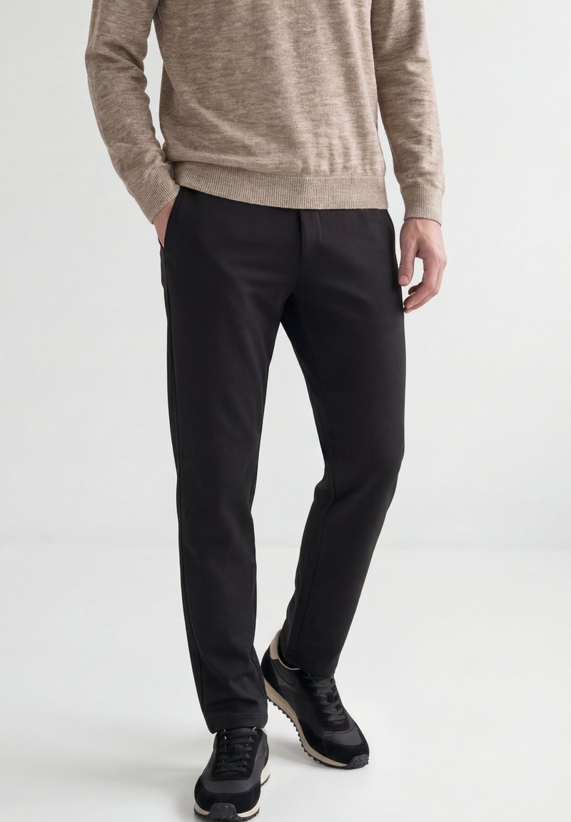 Homme portant un pantalon noir, des baskets noires et un pull beige, une main dans la poche, debout devant un fond clair uni.