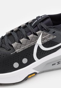 Scarpa da trail running nera Nike con tomaia testurizzata, dettagli riflettenti, sovrapposizioni grigie a motivo e logo Swoosh bianco. Suola Vibram visibile.