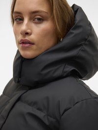 Ung kvinne iført en svart dunjakke med stor høy krage, som ser litt forbi kameraet mot en ensfarget bakgrunn.