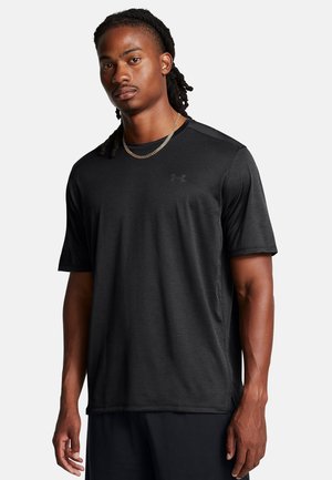 Schwarzes Sport-T-Shirt mit kurzen Ärmeln, glatter Textur, dezentem Schulterdesign und kleinem Logo auf der Brust. Getragen mit einer Halskette.
