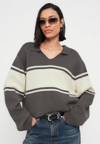 Γυναίκα που φοράει γκρι και λευκή ριγέ μπλούζα, μπλε τζιν με μαύρη ζώνη και γυαλιά ηλίου, στέκεται με τα χέρια στις τσέπες μπροστά σε απλό φόντο.