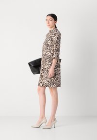 Tijgerprint jurk met driekwart mouwen, beige achtergrond en zwarte accenten. Model houdt een zwarte clutch vast en draagt naakt kleurige hakken.