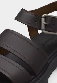 Κοντινή λήψη σκούρου καφέ δερμάτινου σανδαλιού με δύο λουριά και ασημένια αγκράφα στη ρυθμιζόμενη λουρίδα του αστραγάλου.