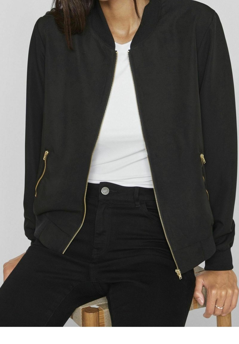 Veste bomber noire avec fermeture éclair sur le devant, col côtelé et poches zippées dorées, fabriquée en tissu lisse. Associée à un haut blanc et un jean noir.