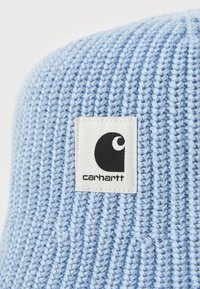 Svijetloplava pletena kapa s rebrastom teksturom i kvadratnom bijelom etiketom s crnim logotipom i imenom brenda "carhartt."
