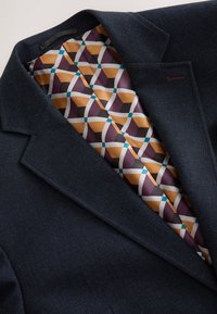 Tmavomodré štruktúrované sako ukazujúce geometrický vzor podšívky v horčicovej, fialovej, bielej, čiernej a tyrkysovej farbe vo vnútri goliera a klopy.